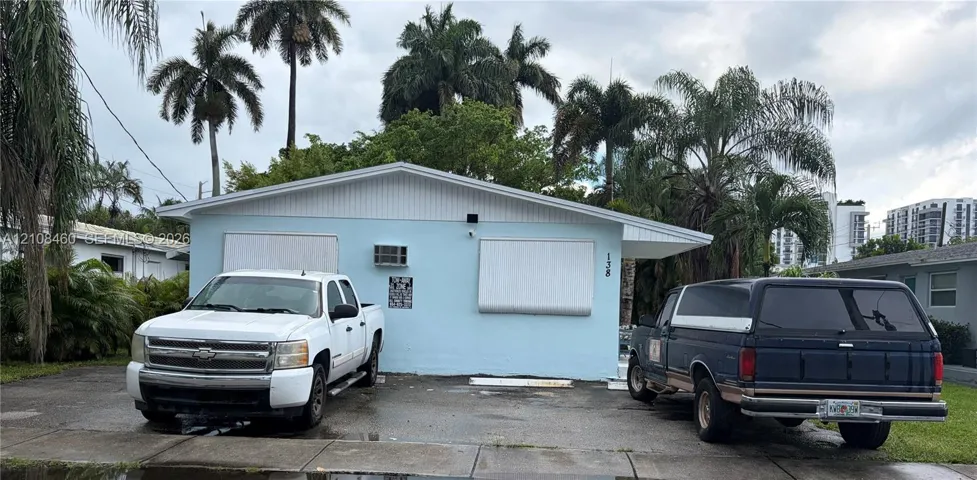 Single-story exterior featuring a light blue facade, a paved parking area, and metal hurricane shutters