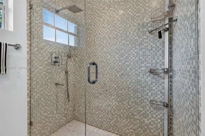 Walk-in shower featuring a frameless glass door, multi-tone mosaic wall tile, overhead rain shower head, handheld shower attachment, and integrated corner shelving