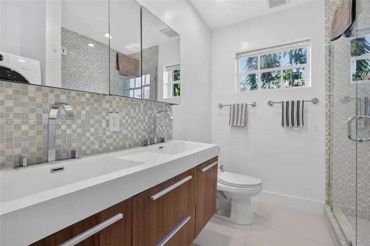 Dual-sink floating vanity with wood-finish cabinetry, mosaic tile backsplash, mirrored medicine cabinets, and glass-enclosed shower