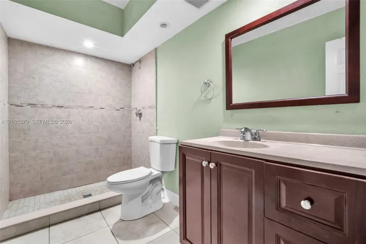 Bathroom featuring a walk-in tiled shower with an accent border, vanity with integrated stone-finish countertop, dark wood-finish cabinetry, and a framed wall-mounted mirror
