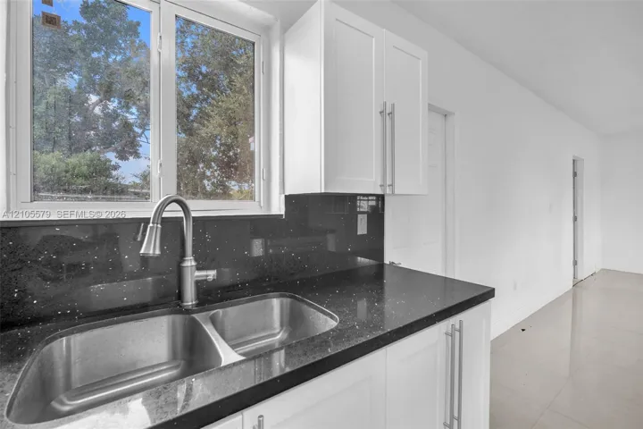 Kitchen area featuring white shaker-style cabinetry, dark stone-finish countertops, stainless steel dual-basin sink, high-arc brushed nickel faucet, and large-format tile flooring