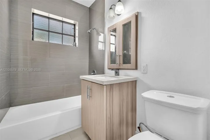 Bathroom featuring a tile-surround bathtub with a rectangular window, light wood-finish vanity cabinet, stone-composite countertop, and a double-door medicine cabinet