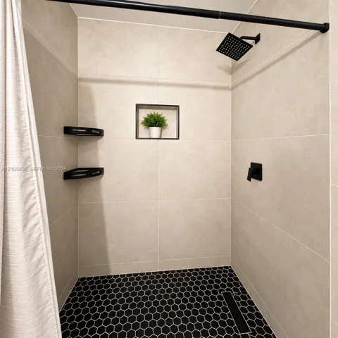 Shower stall featuring large-format beige wall tile, hexagonal black floor tile, matte black rainfall shower head, integrated wall niche, and corner shelving