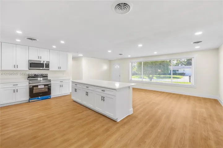 Open-concept kitchen featuring white shaker-style cabinetry, a central island with a solid-surface countertop, stainless steel appliances, recessed lighting, and wood-finish flooring