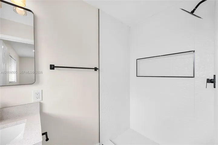 Bathroom featuring a walk-in shower with textured tile surround and built-in recessed shelf, matte black wall-mounted shower head and fixtures, stone-finish countertop with integrated sink, and an arched wall-mounted mirror