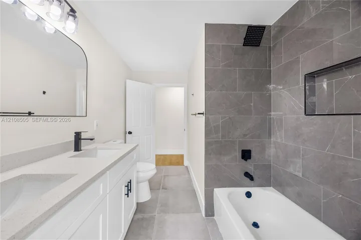 Double vanity with a stone-finish countertop, matte black plumbing fixtures, arched wall mirror, and a bathtub with full-height stone-finish tile surround and built-in recessed niche