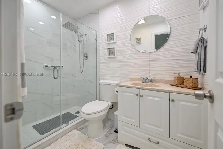 Walk-in shower with floor-to-ceiling stone-patterned tile and rainfall showerhead, white vanity with integrated countertop, and textured accent wall tiling