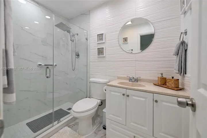Bathroom featuring a glass-enclosed shower with rainfall showerhead, white vanity with integrated basin, stone-surface countertop, and textured wall tile