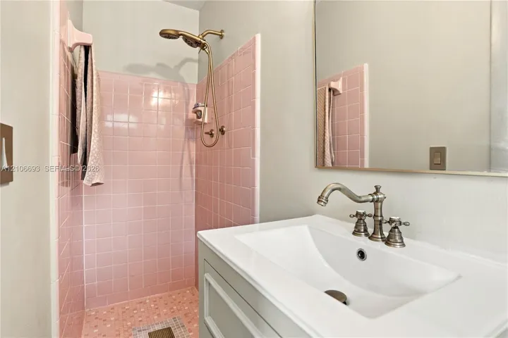 Bathroom featuring square pink tile shower surround, brass-finish handheld shower fixture, rectangular white undermount sink, brass-finish widespread faucet, and large frameless mirror