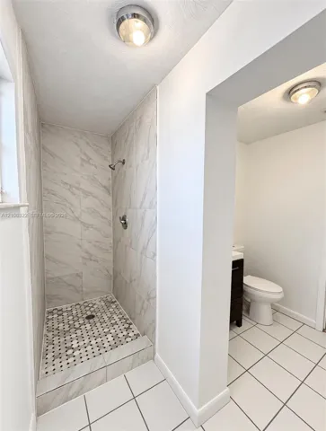 Shower stall featuring large-format light-toned stone-finish wall tile, a decorative hexagonal mosaic tile floor, and a polished metal shower fixture, paired with a separate water closet area featuring large-format white tile flooring