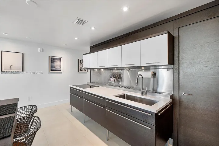 Kitchen featuring white and dark wood-finish cabinetry, light stone-surface countertops, stainless steel backsplash, integrated cooktop, and undermount stainless steel sink