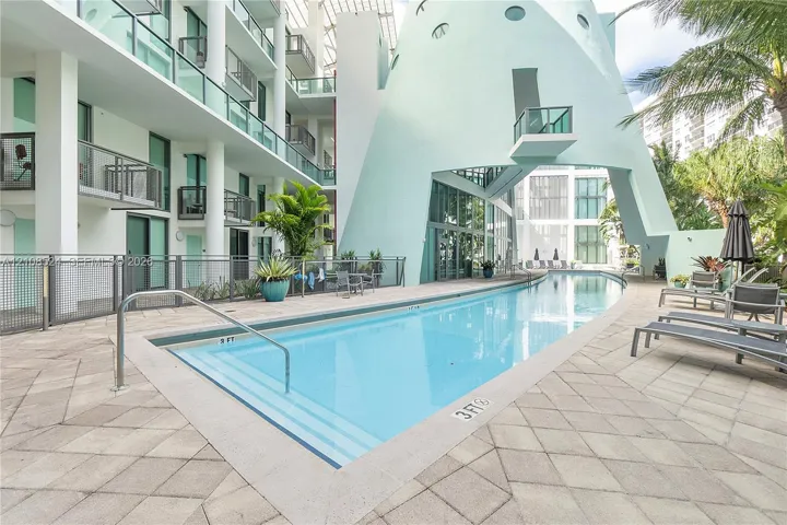 In-ground swimming pool with stepped entry, expansive stone-paver decking, and prominent architectural sculptural facade featuring metal railings and glass balcony enclosures