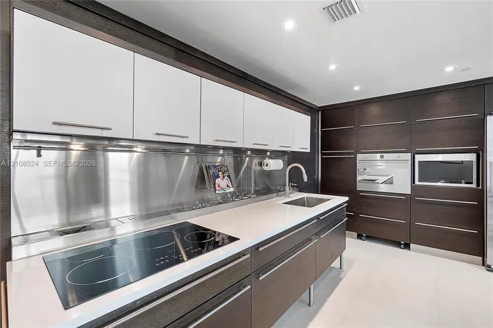 Kitchen featuring white upper cabinetry, dark wood-finish lower cabinetry, metallic backsplash, stone countertops, and built-in stainless steel appliances