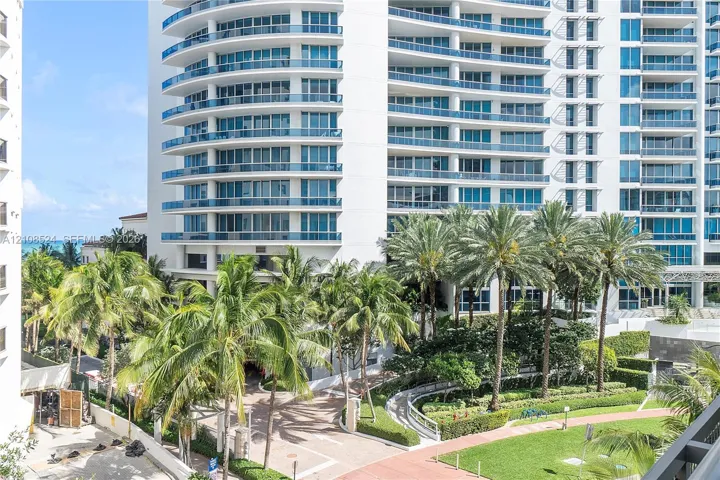 High-rise building exterior featuring curved glass balconies, concrete facade, landscaped grounds with mature palm trees, and paved walkways