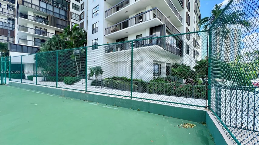 Recreational court with a high-tension perimeter fence, situated on the grounds of a multi-story building featuring private balconies and landscaped walkways
