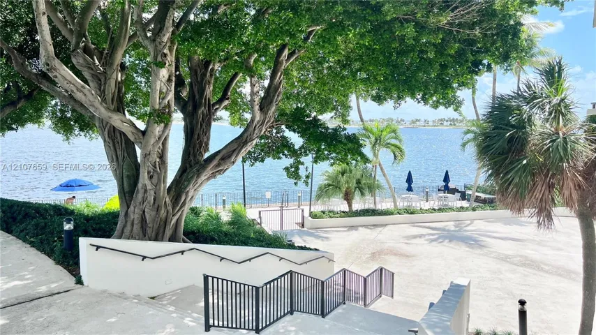 Waterfront grounds featuring mature canopy trees, concrete staircase with black metal railings, and stone-paved promenade