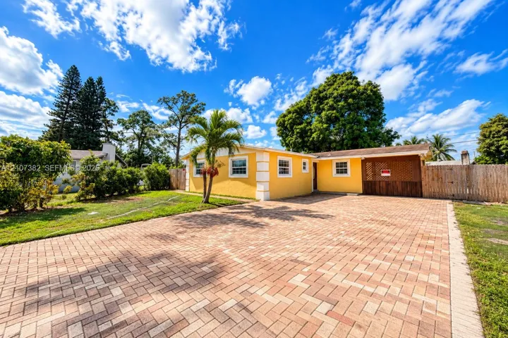 Exterior facade featuring a yellow stucco finish, expansive brick-paver driveway, integrated carport, and wood-latched privacy fencing
