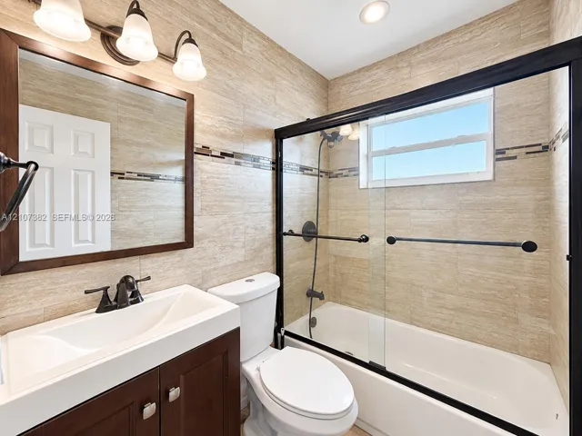 Bathroom featuring full-wall textured tiling, dark wood-finish vanity with a rectangular sink, sliding glass shower door with matte black frame, and coordinated matte black fixtures