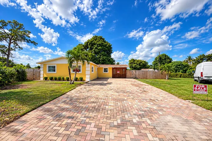 Exterior featuring a multi-tone herringbone paver driveway, yellow stucco facade, white trim accents, and perimeter wood privacy fencing