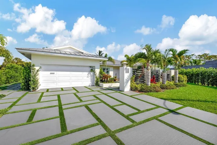 Stucco exterior featuring a dual-car garage, expansive geometric concrete paver driveway with integrated turf ribbons, and mature palm landscaping