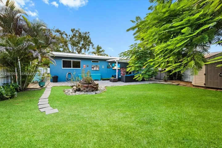 Rear exterior featuring a manicured lawn, stone-paver walkway, rock-bordered garden bed, mature landscaping, and a storage shed