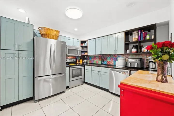 Kitchen featuring light blue cabinetry, stainless steel appliances, patterned tile backsplash, dark countertops, and ceramic tile flooring