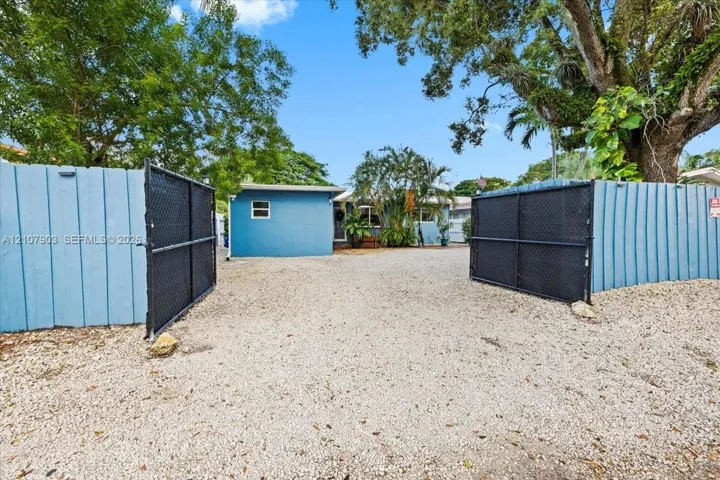Gravel-surfaced driveway entry featuring dual black metal swinging gates and perimeter wood fencing