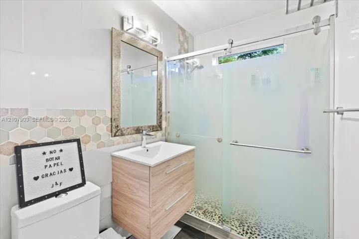 Bathroom featuring a wood-finish vanity with a rectangular sink, walk-in shower with frosted glass sliding doors, stone-tiled shower floor, and hexagonal wall tile backsplash