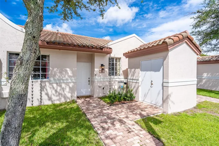 Stucco exterior featuring a clay tile roof, brick paver walkway, white paneled front door, and an attached exterior storage structure