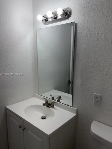 Bathroom vanity featuring a solid-surface countertop with an integrated basin, white cabinetry, polished metal faucet, wall-mounted mirror, and a three-light fixture