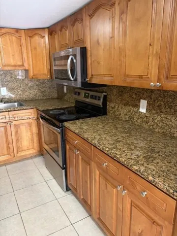 Kitchen featuring arched-panel wood cabinetry, speckled stone countertops, a stainless steel microwave and oven, a dual-basin stainless steel sink, and light-tone ceramic tile flooring
