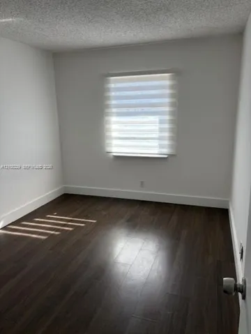 Neutral-toned room featuring dark wood-finish flooring, a single-pane window with zebra-style roller shades, and a textured ceiling finish