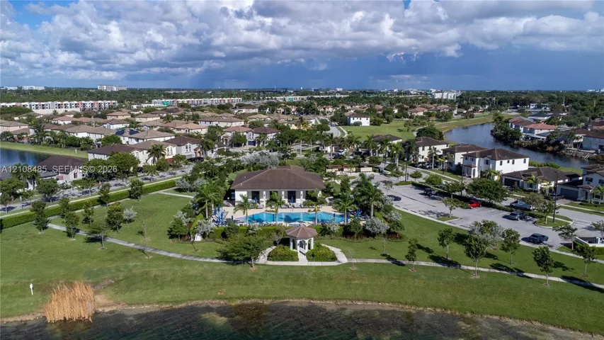 Aerial perspective of a community facility featuring a swimming pool, covered gazebo, surrounding landscaped grounds, and a paved parking area