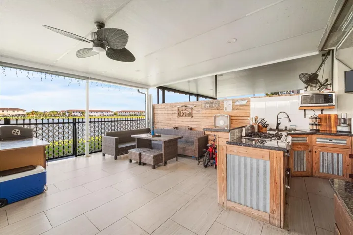 Covered patio featuring large-format tile flooring, a ceiling fan, and a built-in outdoor kitchen with stone-finish countertops, wood cabinetry with metal paneling, and a high-arch faucet