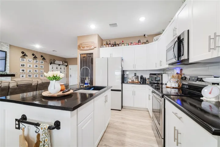 Kitchen featuring white cabinetry, dark countertops, an island with an integrated sink, stainless steel appliances, and wood-finish flooring