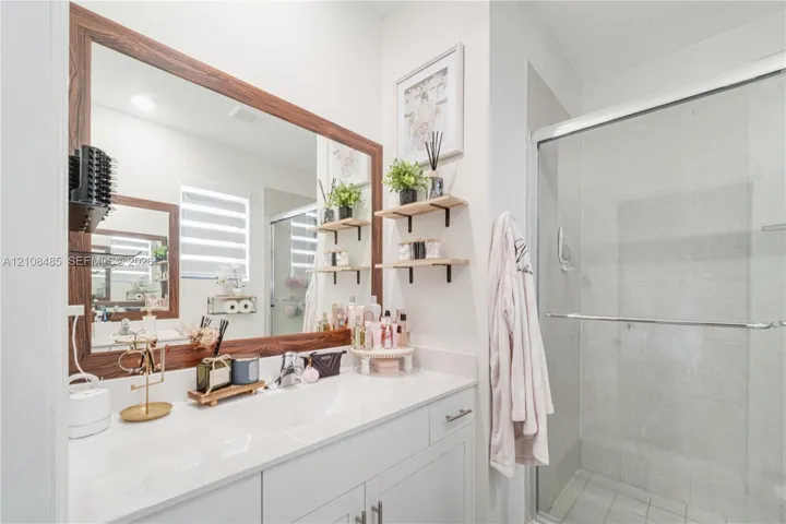 Vanity featuring a white countertop with an integrated basin, wood-frame mirror, glass-enclosed shower, and wall-mounted shelving