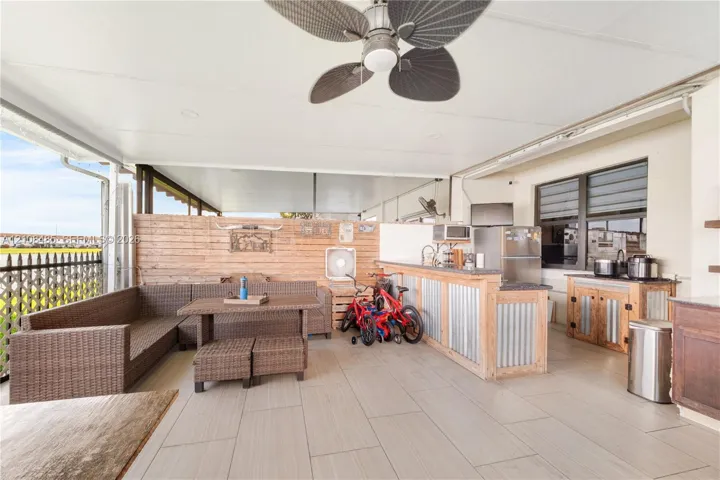 Covered outdoor patio featuring light-toned tile flooring, built-in bar with metal siding, horizontal wood-slat privacy wall, ceiling fan, and louvered windows