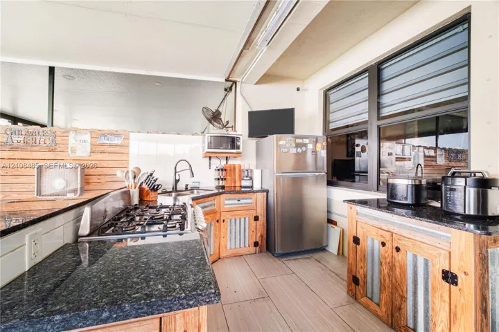 Outdoor kitchen featuring dark stone countertops, custom wood cabinetry with corrugated metal detailing, built-in stainless steel gas range, and large-format tile flooring