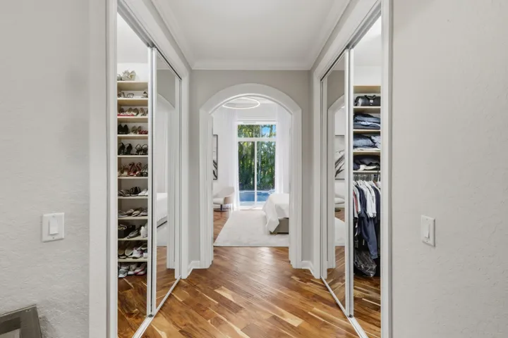 His and Hers walk-in wardrobe featuring mirrored sliding doors, custom integrated shelving, wood-finish flooring, and a decorative arched entryway