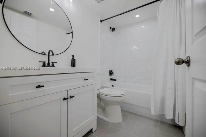 White vanity featuring matte black hardware and a circular mirror, paired with a shower-tub combination finished in white subway tile and matte black fixtures, complemented by large-format tile flooring