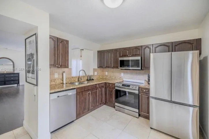 Kitchen featuring dark wood cabinetry, stone-surface countertops, tile backsplash, and stainless steel appliances including a refrigerator, range, microwave, and dishwasher