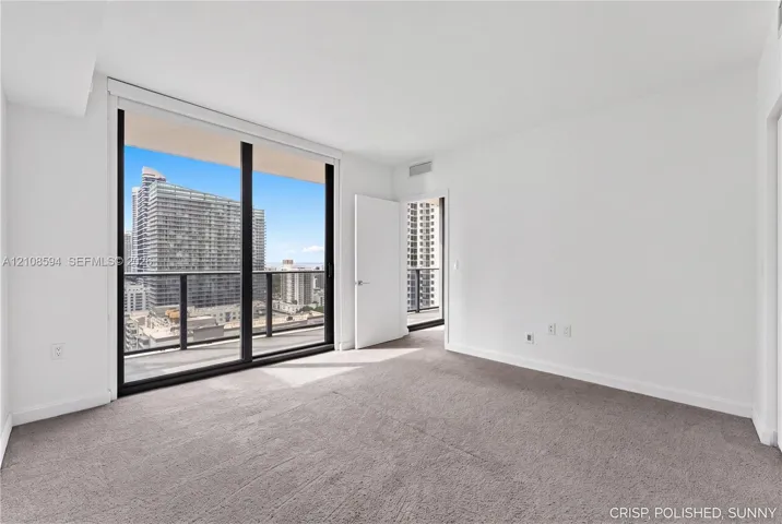 Floor-to-ceiling glass sliding doors, private balcony with metal railing, carpeted flooring, and urban cityscape scenery