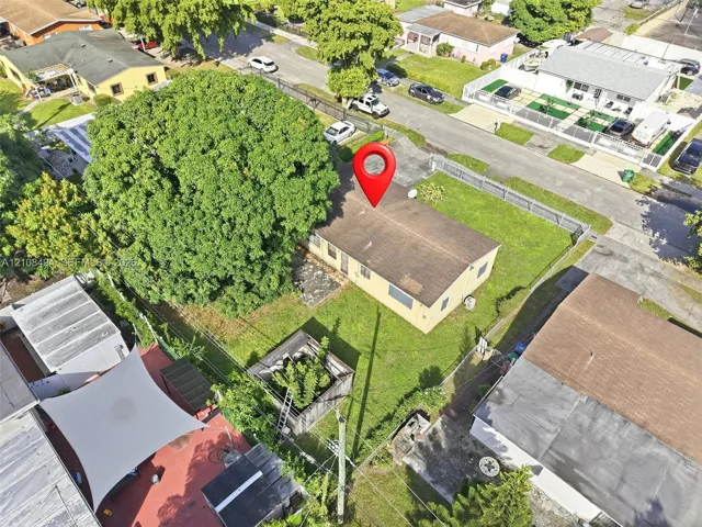 Aerial perspective of a detached single-story residence featuring a shingled roof, exterior masonry siding, a fenced perimeter, and mature canopy trees on a grass-covered lot