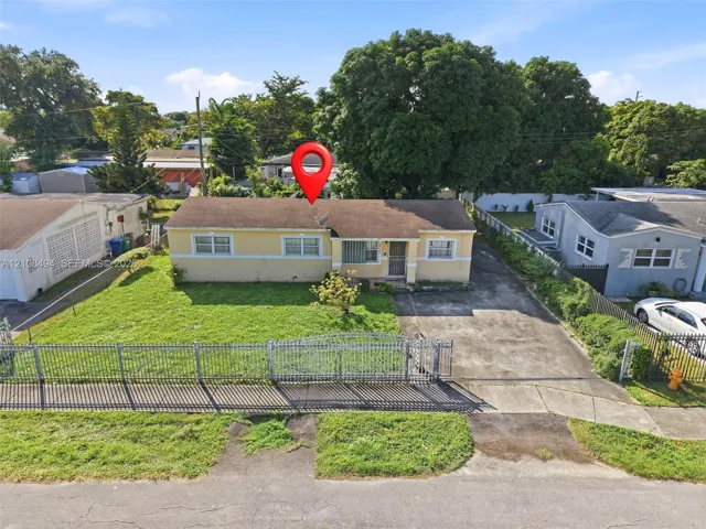 Single-story residence featuring a low-slope roof, stucco exterior, paved driveway, metal perimeter fencing, and mature trees