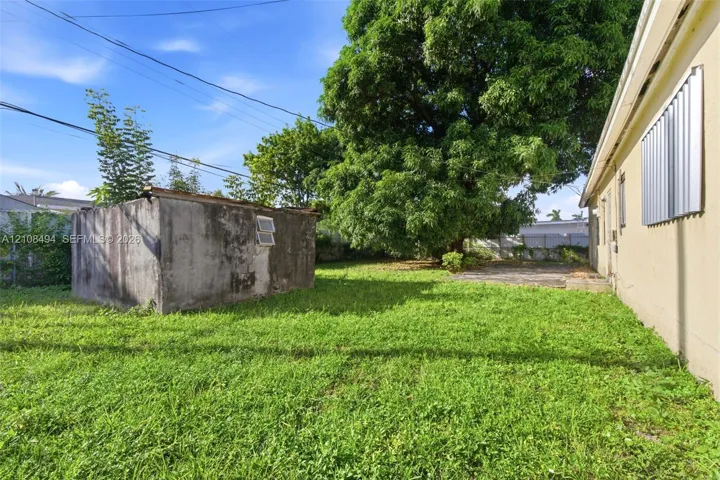 Rear yard featuring a grass lawn, mature tree, masonry outbuilding, and concrete patio area