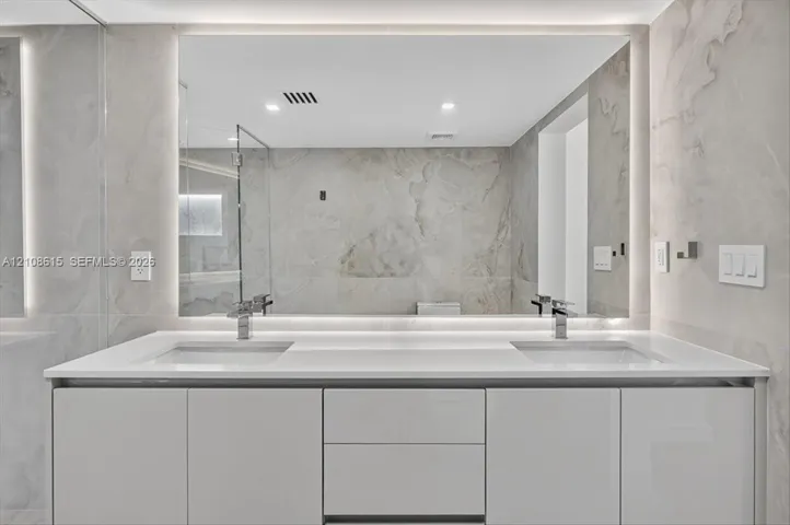 Modern dual-sink vanity featuring a solid-surface countertop, high-gloss cabinetry, integrated LED-backlit mirror, and polished metal fixtures against full-height stone-finish wall tile