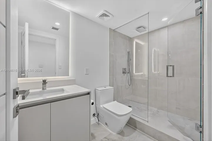 Modern bathroom featuring a floating vanity with a light-toned countertop, a seamless walk-in shower with a glass enclosure, large-format tile flooring, and backlit wall mirror
