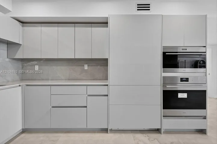 Modern kitchen featuring flat-panel cabinetry, stone-finish backsplash, neutral-toned countertops, and integrated stainless steel wall ovens