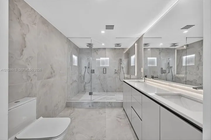 Dual-sink vanity with high-gloss cabinetry and solid surface countertop, walk-in glass-enclosed shower featuring dual rainfall showerheads and recessed wall niches, and floor-to-ceiling large-format tile surround