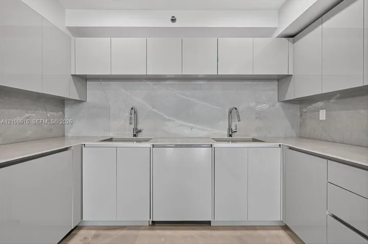 Glossy cabinetry, stone slab backsplash, dual undermount sinks, gooseneck faucets, and light-toned countertop surfaces